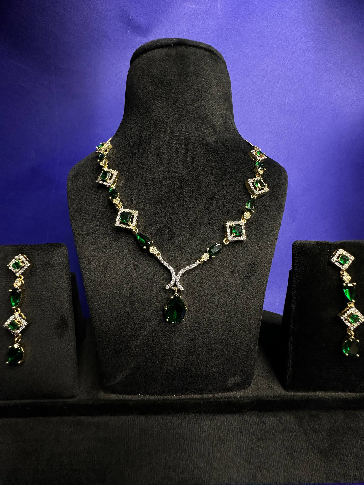 A gold and green gemstone jewelry set displayed on a black velvet mannequin bust, featuring a necklace with a teardrop pendant and matching drop earrings, set against a purple background.