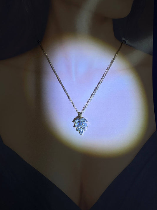 A delicate gold chain necklace with a leaf-shaped pendant set with clear stones, shown worn on a model's neck under a dramatic spotlight.