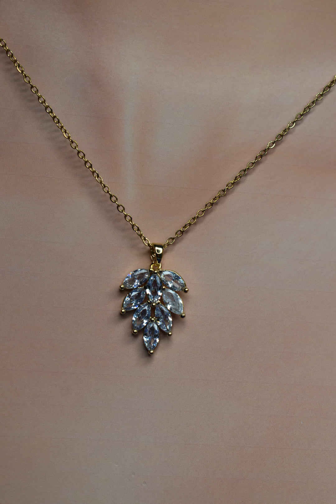 A gold chain necklace with a leaf-shaped pendant featuring marquise-cut clear crystals, displayed on a soft beige background.