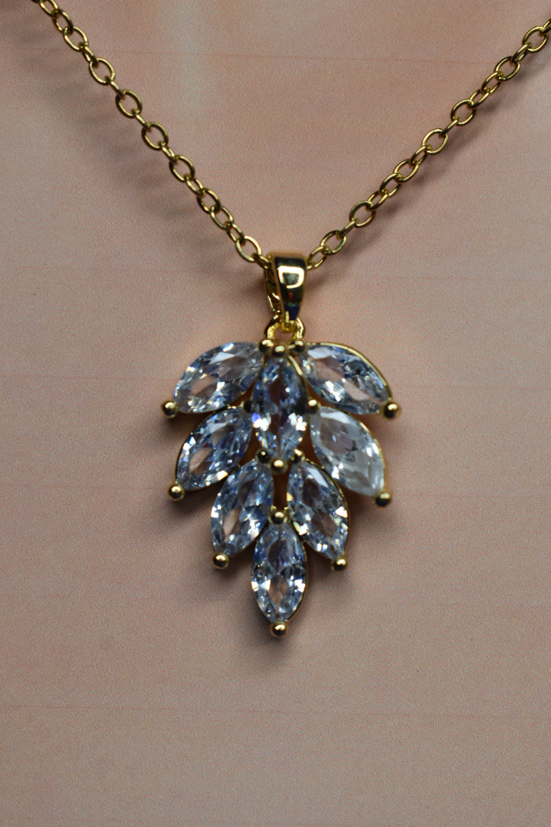 Gold chain necklace with a leaf-shaped pendant featuring marquise-cut light blue crystals on a soft pink background.
