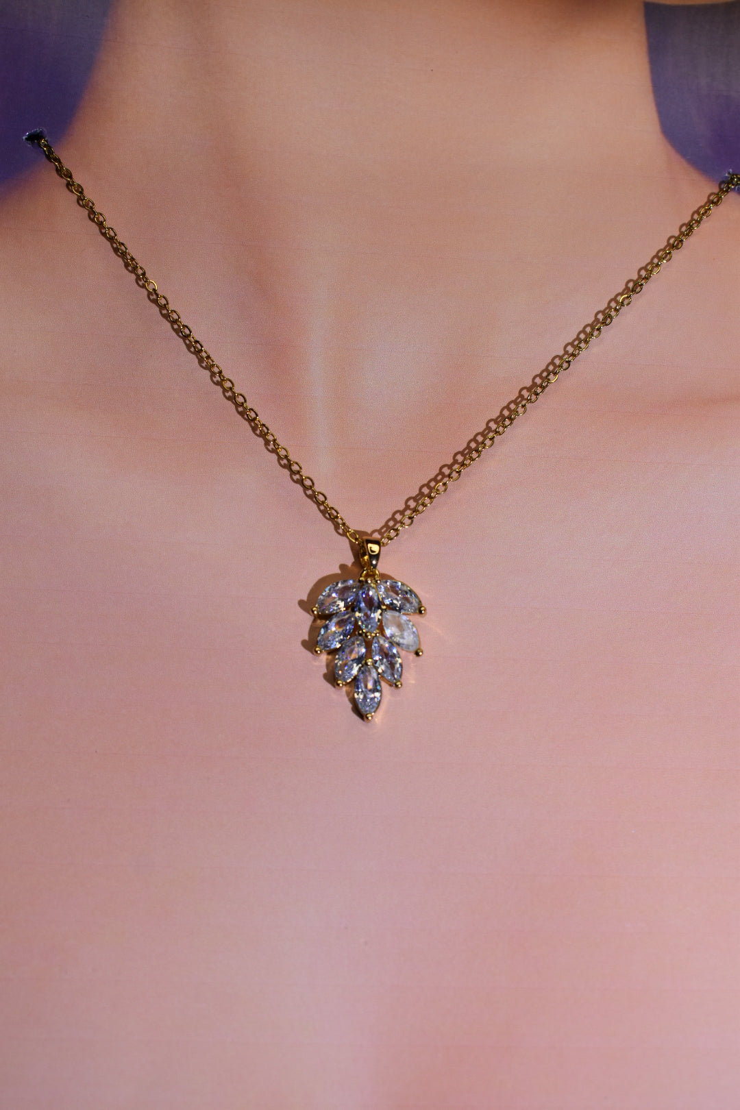 A gold chain necklace with a leaf-shaped pendant set with clear gemstones, shown worn on a model's neck against a soft pink background.