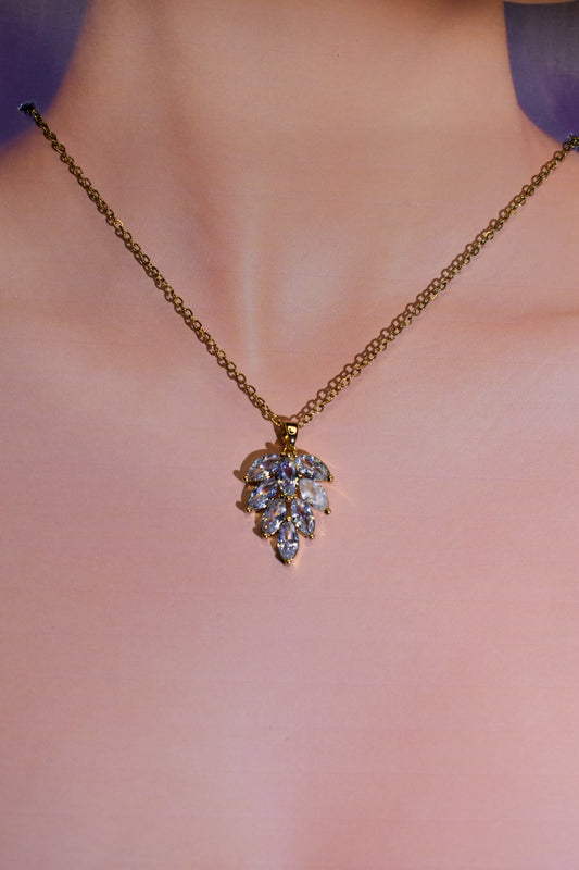 A gold chain necklace with a leaf-shaped pendant set with clear gemstones, shown worn on a model's neck against a soft pink background.