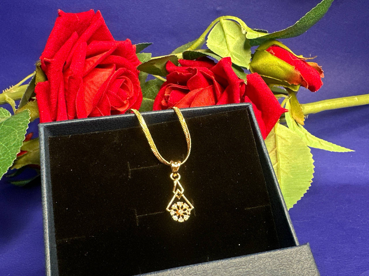 A gold pendant necklace with a geometric starburst design hangs in an open black jewelry box, styled with red roses on a purple background.