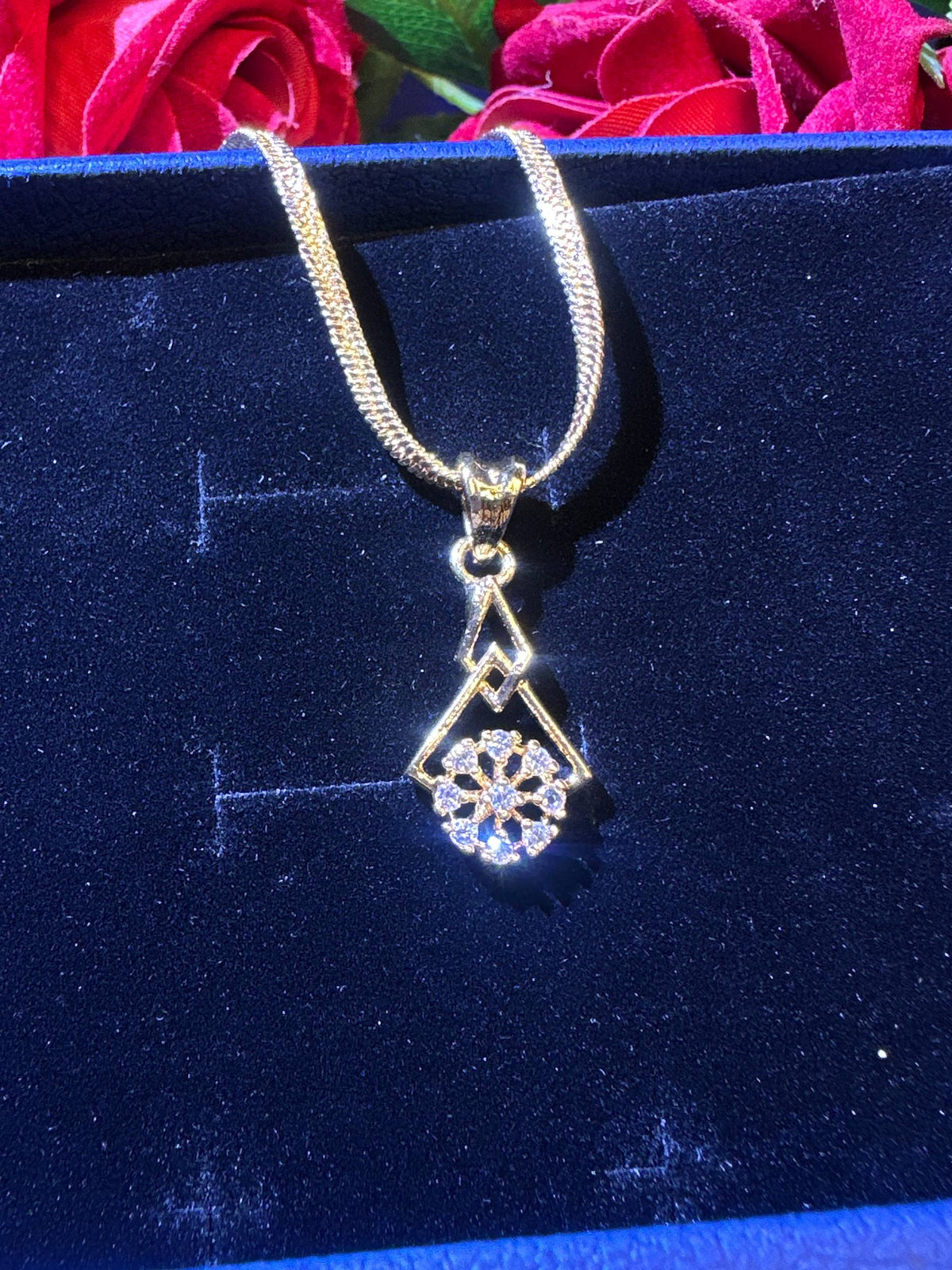 A gold chain necklace with a geometric pendant featuring a floral snowflake design, displayed on a dark blue velvet jewelry box with red roses in the background.