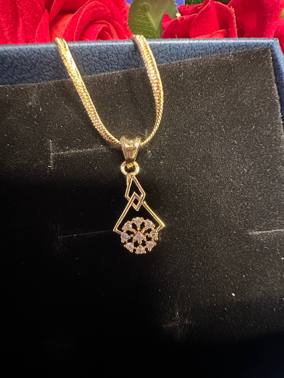 A gold pendant necklace with a geometric mountain and floral design, resting on a black jewelry box with red roses in the background.