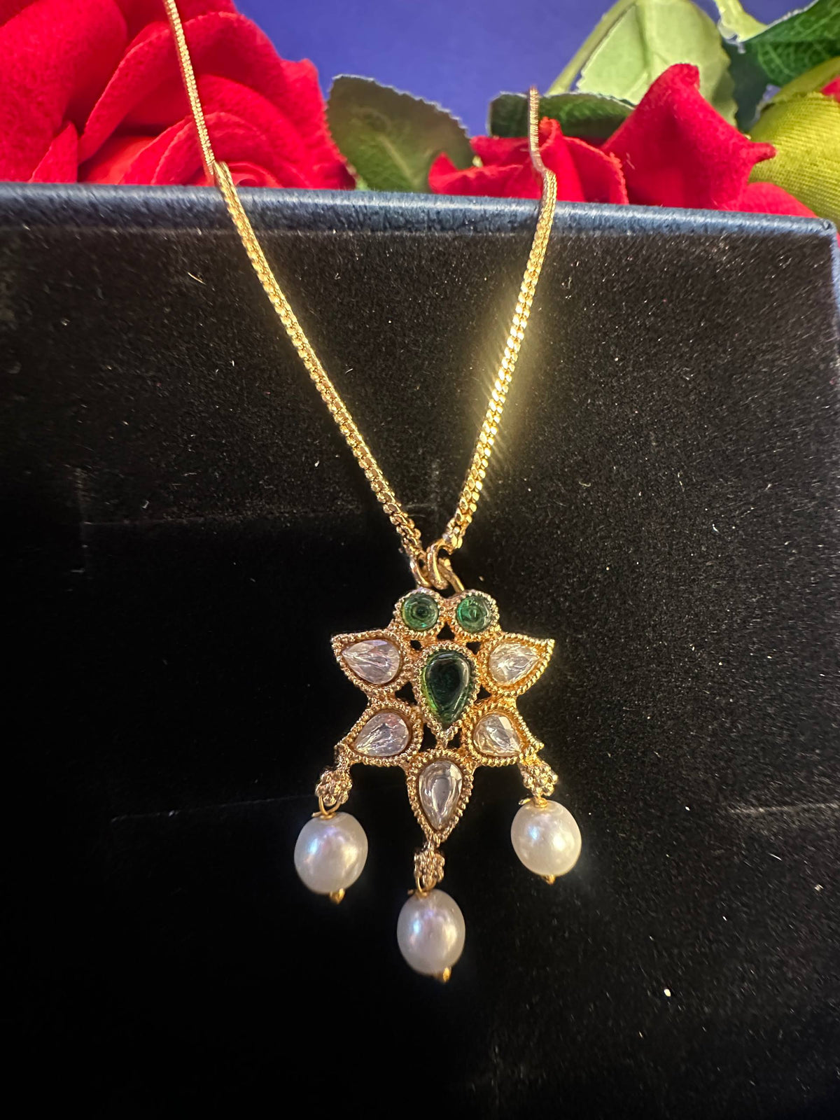 Gold pendant necklace with green and white stones and three dangling pearls, displayed on a black surface with red roses in the background.