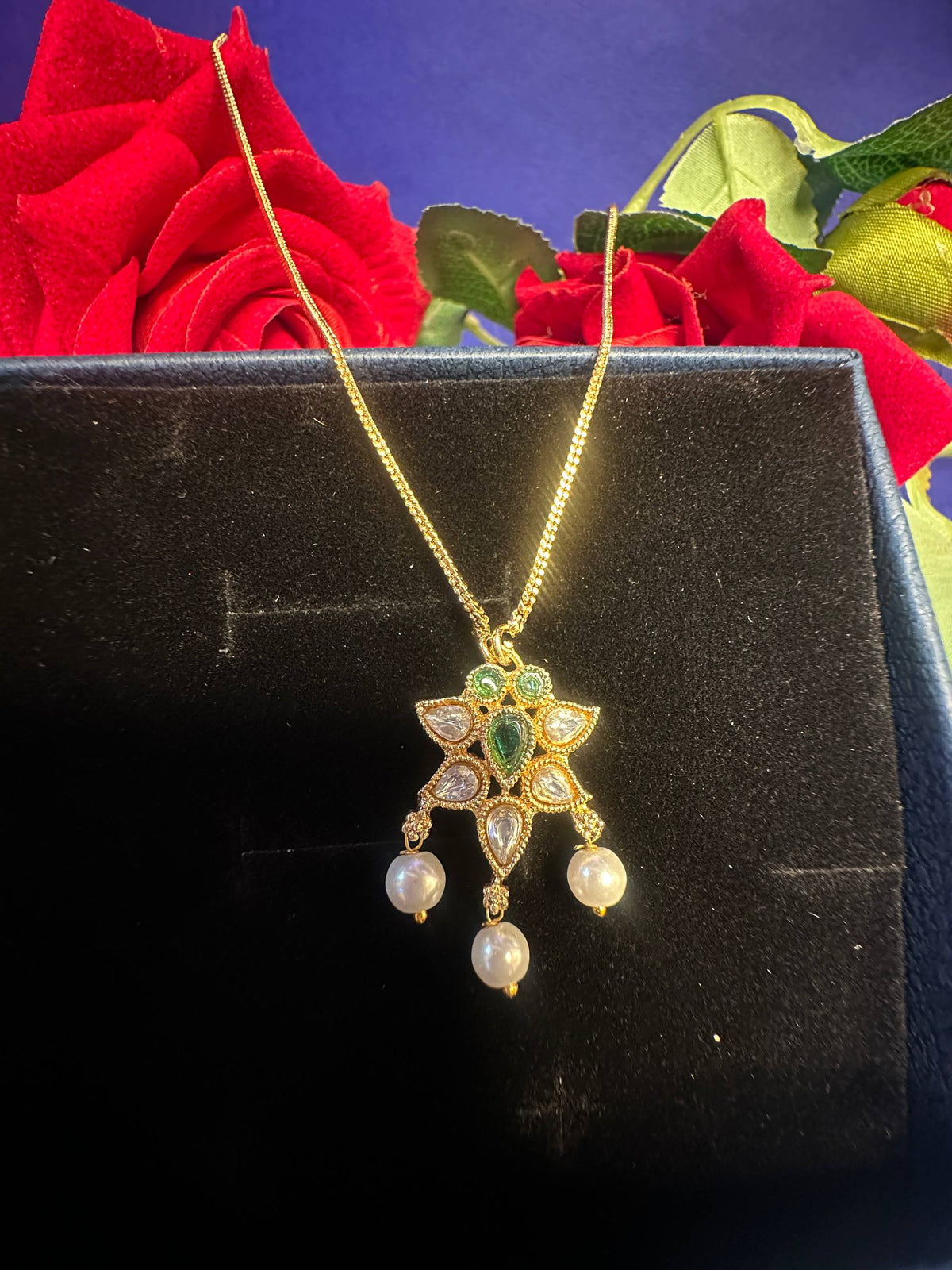 Gold pendant necklace with green and white stones and pearl drops, displayed on a black box with red roses in the background.