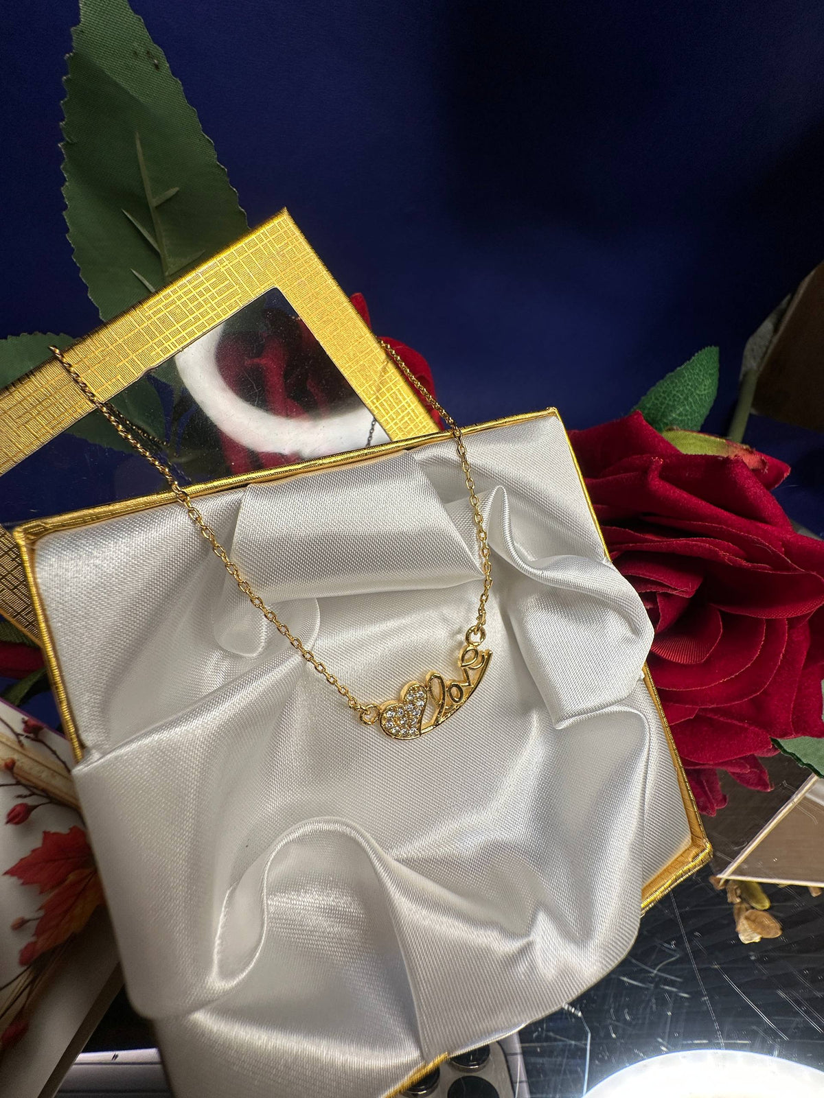 A gold necklace with a heart-shaped pendant displayed on a white satin ribbon draped over a gold box, surrounded by red roses and a dark blue background.