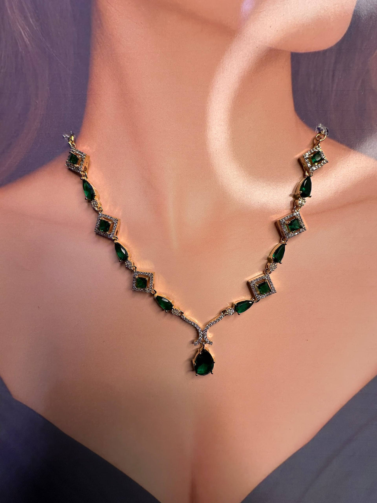 A close-up of a woman's neck and upper chest wearing a gold-tone necklace with emerald-cut green gemstones and small clear accent stones.