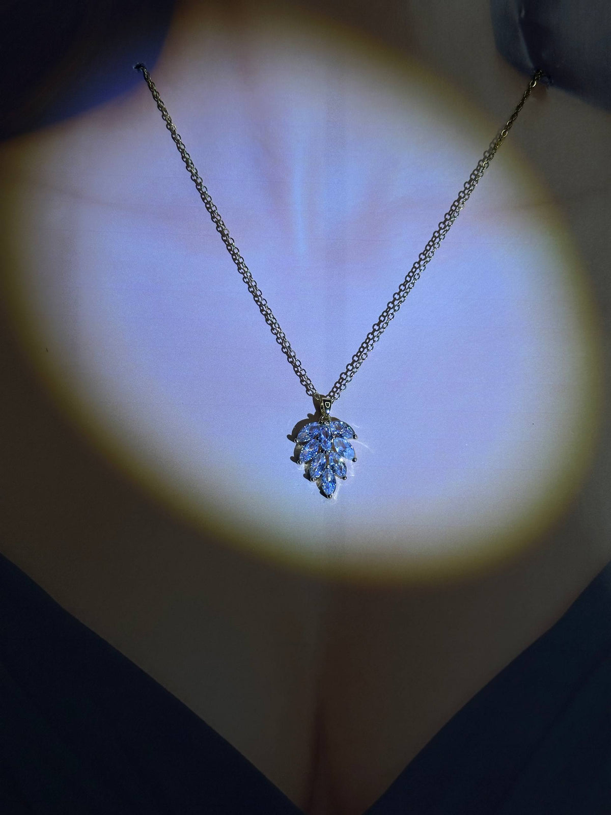 A gold chain necklace with a leaf-shaped pendant set with clear stones, shown worn on a model's neck under a dramatic spotlight.